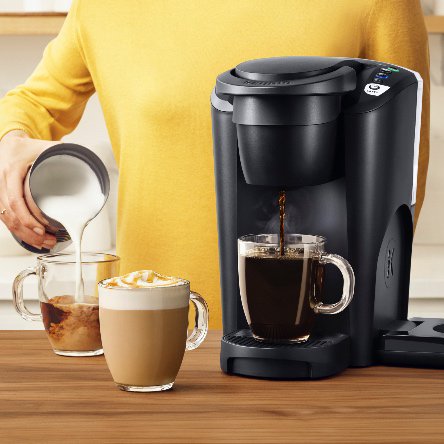 A woman pouring frothed milk into a cup of espresso next to a Keurig K-latte brewer.